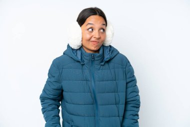 Young hispanic woman wearing a winter earmuffs isolated on white background making doubts gesture while lifting the shoulders