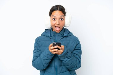 Young hispanic woman wearing a winter earmuffs isolated on white background surprised and sending a message