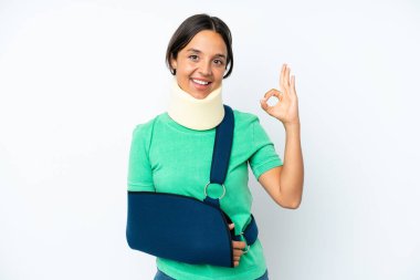 Young hispanic woman wearing a neck brace and sling isolated on white background showing ok sign with fingers
