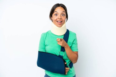 Young hispanic woman wearing a neck brace and sling isolated on white background surprised and pointing front