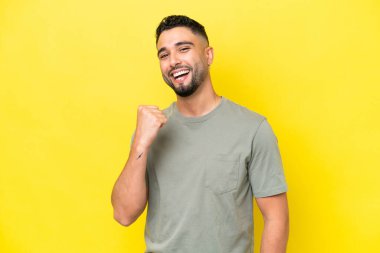Sarı arka planda izole edilmiş genç Arap yakışıklı bir adam zaferi kutluyor.