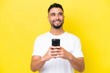 Young Arab handsome man isolated on yellow background using mobile phone and looking up