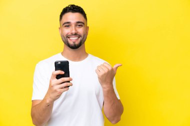 Young Arab handsome man isolated on yellow background using mobile phone and pointing to the lateral