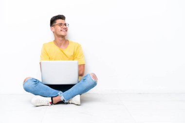 Young man sitting on the floor isolated on white background looking up while smiling