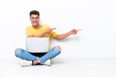 Genç adam, beyaz arka planda izole bir şekilde yerde oturuyor. Parmağını kenarı gösteriyor ve bir ürün sunuyor.