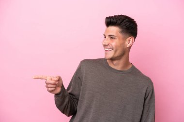 Pembe arka planda izole edilmiş genç beyaz bir adam parmak göstererek bir ürün sunuyor.