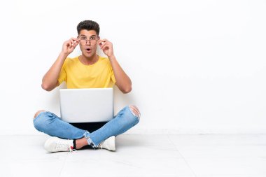 Genç adam beyaz arka planda gözlüklü ve şaşırmış bir şekilde yerde oturuyor.