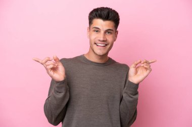 Pembe arka planda izole edilmiş genç beyaz adam parmak uçlarını işaret ediyor ve mutlu.