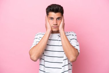 Pembe arka planda izole edilmiş genç beyaz adam hayal kırıklığına uğramış ve kulaklarını kapatmış.