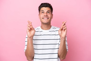 Pembe arka planda izole edilmiş genç beyaz adam parmaklarını çapraz yapıp en iyisini diliyor.