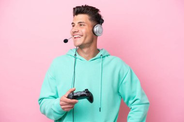 Young handsome caucasian man playing with a video game controller isolated on pink background posing with arms at hip and smiling