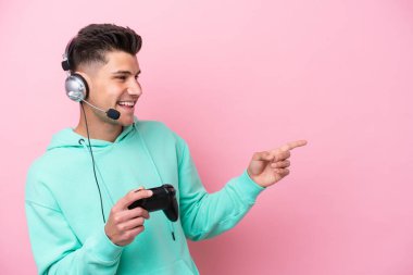 Young handsome caucasian man playing with a video game controller isolated on pink background pointing finger to the side and presenting a product