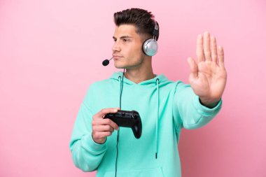 Young handsome caucasian man playing with a video game controller isolated on pink background making stop gesture and disappointed