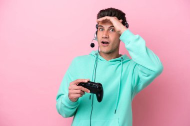 Young handsome caucasian man playing with a video game controller isolated on pink background doing surprise gesture while looking to the side