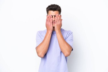Beyaz arka planda izole edilmiş beyaz tenli genç adam gözlerini elleriyle kapatıyor.