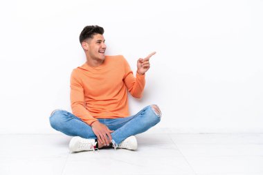 Genç adam, beyaz arka planda izole bir şekilde yerde oturuyor. Parmağını kenarı gösteriyor ve bir ürün sunuyor.