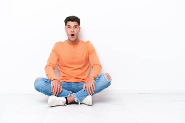 Yerde oturan genç adam, beyaz arka planda, şaşırmış bir yüz ifadesiyle izole edilmiş.