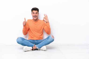 Yerde oturan genç adam beyaz arka planda izole bir şekilde tamam işareti ve baş parmak işareti gösteriyor.