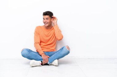 Genç adam beyaz arka planda izole bir şekilde yerde oturuyor. Kulağına elini sürerek bir şeyler dinliyor.