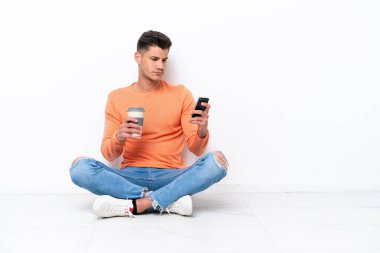 Genç adam beyaz arka planda izole bir şekilde yerde oturuyor. Elinde kahve ve cep telefonu var.