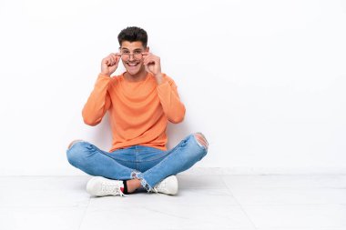 Genç adam beyaz arka planda gözlüklü ve şaşırmış bir şekilde yerde oturuyor.