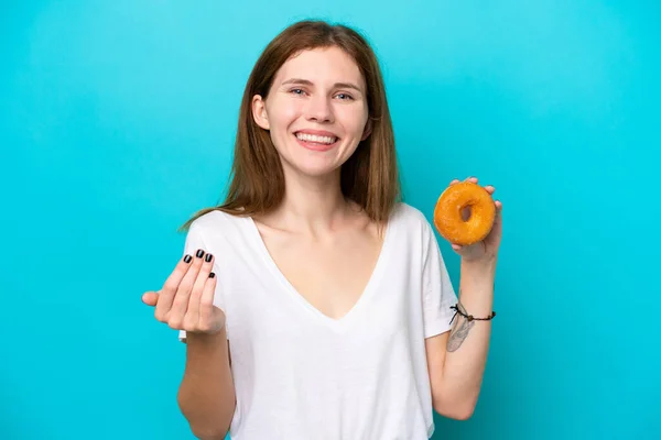 Young English woman holding a donut over isolated blue background inviting to come with hand. Happy that you came