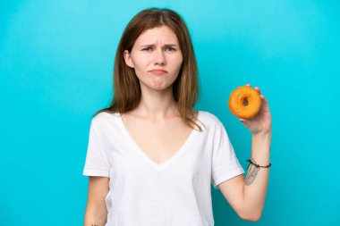 Soyutlanmış mavi arkaplanda üzgün bir ifadeyle donut tutan genç bir İngiliz kadın.