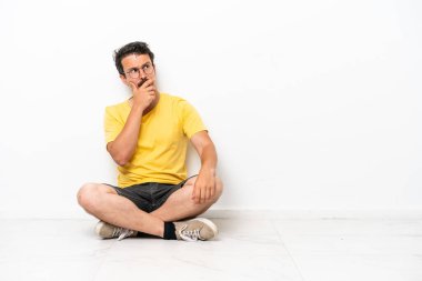 Beyaz arka planda izole bir şekilde yerde oturan genç beyaz adamın şüpheleri ve şaşkın bir yüz ifadesi var.