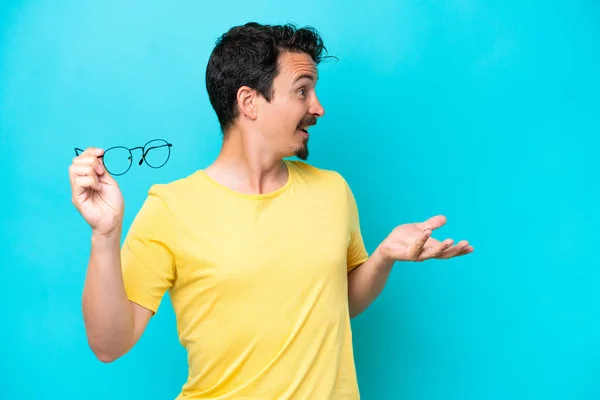 Young Caucasian Man Holding Glasses Isolated Blue Background Surprise Facial — Stok Foto