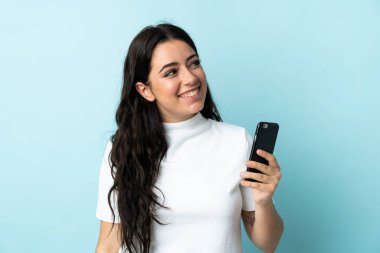 Young woman using mobile phone isolated on blue background looking up while smiling