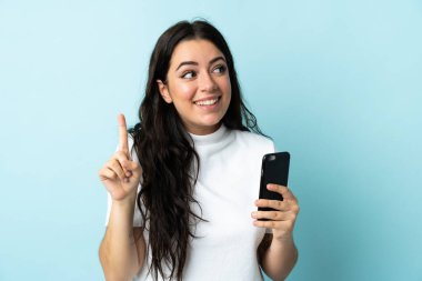 Young woman using mobile phone isolated on blue background intending to realizes the solution while lifting a finger up