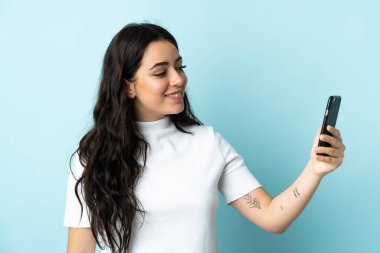 Mavi arka planda izole edilmiş ve mutlu bir ifadeyle cep telefonu kullanan genç bir kadın.