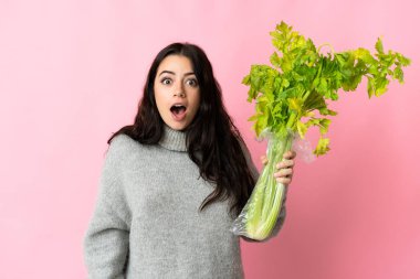 Young caucasian woman holding a celery isolated on blue background with surprise and shocked facial expression