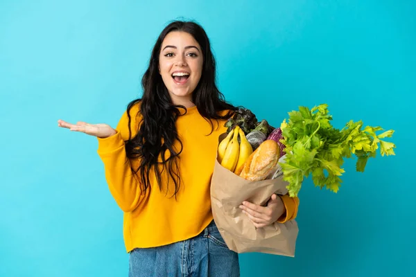 Young Woman Holding Grocery Shopping Bag Isolated Blue Background Shocked — стоковое фото