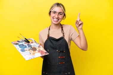 Young artist caucasian woman holding a palette isolated  on yellow background pointing up a great idea