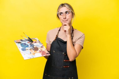 Young artist caucasian woman holding a palette isolated  on yellow background having doubts and thinking
