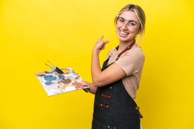 Young artist caucasian woman holding a palette isolated  on yellow background pointing back