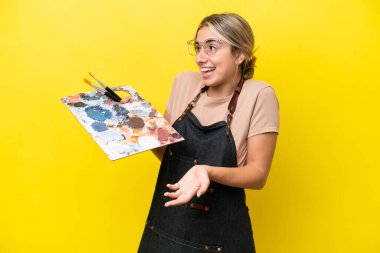 Young artist caucasian woman holding a palette isolated  on yellow background with surprise expression while looking side