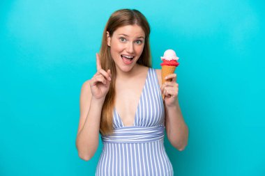 Mavi arka planda izole edilmiş, mayo giymiş, dondurma yiyen beyaz bir kadın harika bir fikre işaret ediyor.