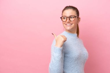 Pembe arka planda izole edilmiş genç beyaz kadın bir ürünü sunmak için kenarı işaret ediyor.