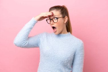 Pembe arka planda izole edilmiş genç beyaz kadın yan tarafa bakarken sürpriz jest yapıyor.