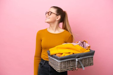 Pembe arka planda izole edilmiş çamaşır sepetini tutan beyaz bir kadın yan pozisyonda gülüyor.