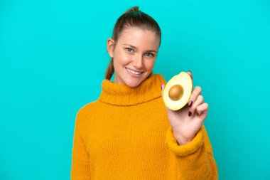 Avokado tutan beyaz bir kadın mavi arka planda mutlu bir ifadeyle izole edilmiş.