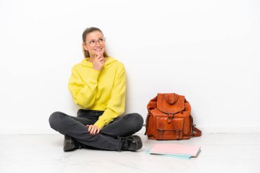 Beyaz arka planda zemin katta oturan beyaz tenli genç bayan yan tarafa bakıyor.