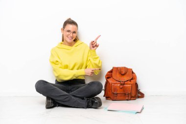 Beyaz arka planda beyaz bir zeminde oturan beyaz tenli genç bir kadın parmak göstererek bir ürün sunuyor.