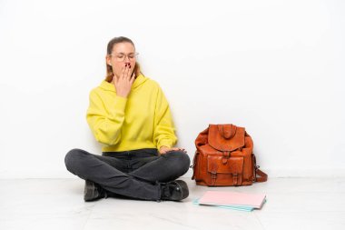 Beyaz arka planda tek başına oturan beyaz tenli genç bir kadın esniyor ve ağzını eliyle kapatıyor.