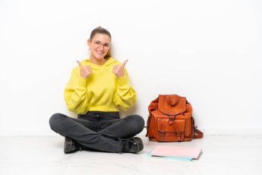 Beyaz arka planda beyaz bir zeminde oturan beyaz tenli genç bir kadın. Baş parmakları havada ve gülümsüyor.