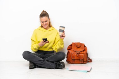 Beyaz arka planda bir katta oturan beyaz tenli genç bir kadın kahve alıp götürmek için elinde bir cep telefonu var.