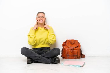 Beyaz arka planda beyaz bir zeminde oturan beyaz bir kadın mutlu ve hoş bir ifadeyle gülümsüyor.