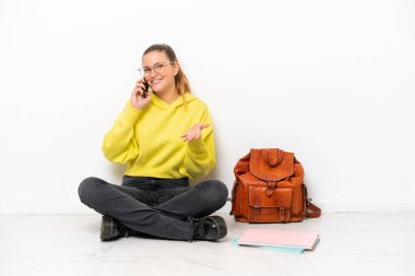 Beyaz arka planda bir katta oturan beyaz tenli genç bir kadın cep telefonuyla sohbet ediyor.